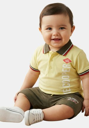 Bébé souriant assis, portant un polo jaune avec un col foncé et un short, ainsi que des baskets blanches sur un fond clair uni.