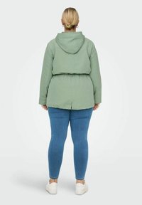 Žalia gobtuvu striukė su raišteliu juosmenyje, suklijuota tekstūra ir šoniniais plyšiais, derinama su priglundančiomis mėlynomis džinsomis ir baltomis sportbatėmis.