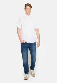 Camisa blanca de manga corta con botones y bolsillo en el pecho, combinada con vaqueros azules y zapatillas beige. Corte recto, ajuste casual.
