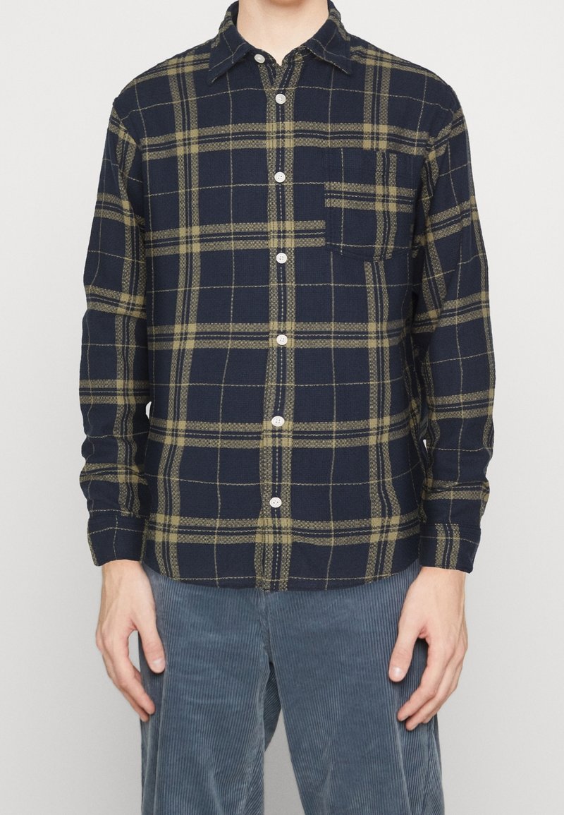 Camisa a cuadros en tonos navy con acentos verdes, diseño de botones y bolsillo en el pecho izquierdo. Tela texturizada con mangas largas y detalle de puños con botones.