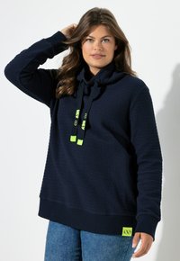 Sudadera con capucha azul marino de tela texturizada, cuello tipo capucha holgada y detalles de cordón en verde neón. Presenta un dobladillo de color sólido y una etiqueta con el logo.
