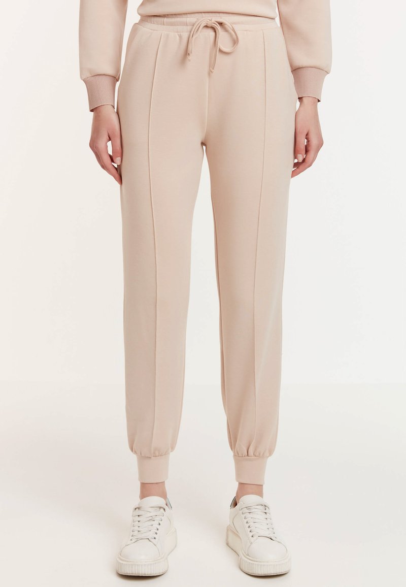 Beige joggers met elastische boorden en een tailleband met trekkoord. Kenmerkt zich door twee verticale naden aan de voorkant. Draagt een paar witte sneakers.
