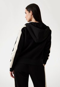 Crna hoodie s bijelim bočnim trakama, koja ima kapu i rebraste manžete. Izrađena od mekanog materijala, prikazuje opušteni kroj i minimalistički dizajn.