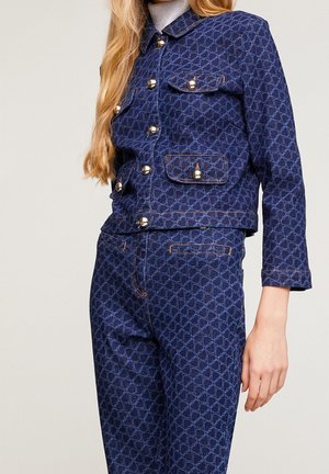 Vrouw die een bijpassend donkerblauw patroon denimjack en hoog getailleerde jeans draagt met gouden knoopdetails en voorzakken, staand tegen een effen achtergrond.