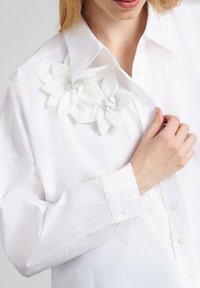 Camisa blanca de manga larga, con un broche floral decorativo blanco en el lado izquierdo del cuello, hecho de tela texturizada. Los botones son visibles.