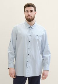 Hellblaues Button-Up-Hemd mit strukturiertem Muster, schwarzen Knöpfen und einer Brusttasche. Verfügt über lange Ärmel mit geknöpften Manschetten.