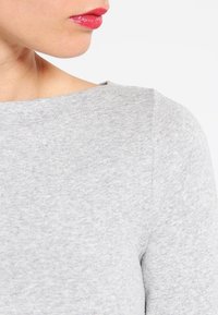 Närbild av en persons nedre ansikte och nacke, klädd i ljusgrå tygkläder med båtutskärning, läpparna är något isär med rosa gloss.