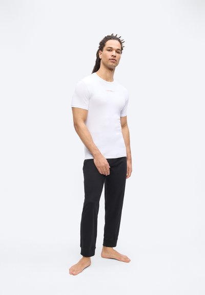 Άνδρας με dreadlocks φοράει εφαρμοστή λευκή κοντομάνικη πουκάμισα και μαύρο παντελόνι, στέκεται ξυπόλητος μπροστά σε ένα απλό λευκό φόντο.