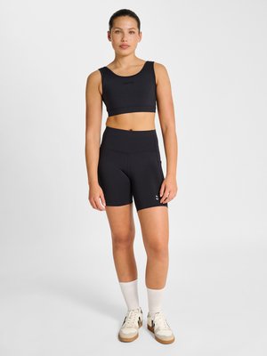 Ung kvinde stående iført en sort crop top, højtaljede sorte biker-shorts, hvide sokker og beige sneakers på hvid baggrund.