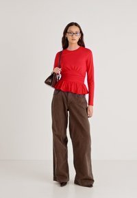 Rød peplum-topp med lange ermer kombinert med løse brune bukser, med teksturert stoff og en skulderveske, plassert mot en nøytral bakgrunn.