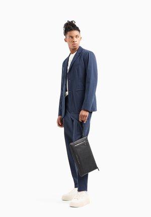 Completo due pezzi di colore blu navy con finitura texturizzata, abbinato a sneakers bianche. Tiene in mano una borsa nera con texture, dotata di zip e manico.