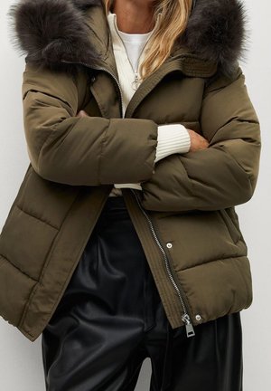 Person wearing an olive green puffer jacket with fur hood, white sweater, and black leather pants, arms crossed in front of a white background.