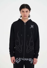 Sudadera negra con cremallera y diseño de llamas de tachuelas plateadas, con capucha ajustable y puños de canalé. Combinada con pantalones de chándal negros con gráfico.
