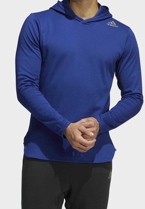 Mężczyzna w niebieskiej bluzie z kapturem z długim rękawem i czarnych spodniach z logo Adidas, stoi z rękami złożonymi z przodu.
