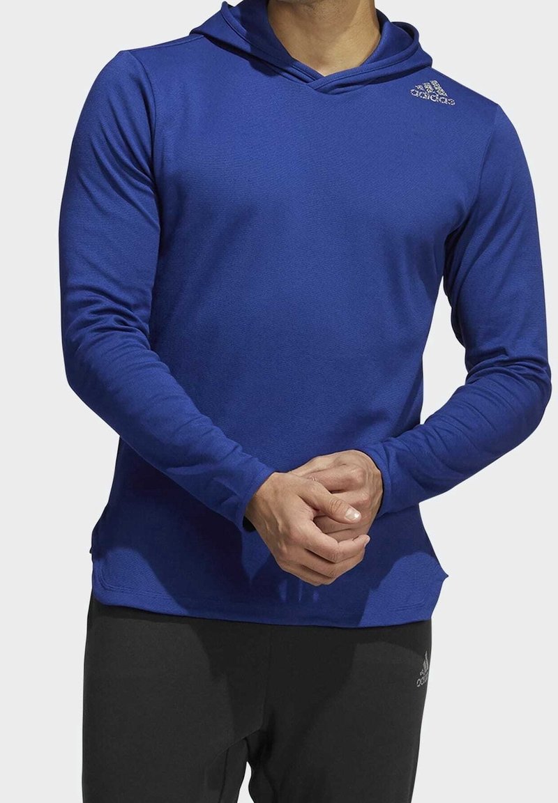 Homme portant un sweat à capuche bleu à manches longues et un pantalon noir avec des logos Adidas, debout les mains jointes devant.