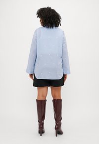 Gaiši zila garām piedurknēm krekls ar izšūtām baltām ziedu rakstiem, aizmugurējā izdiluma pleca šuve, izliekts apakšmalas galu, valkāta ar melnu svārku un bordo ceļgalu augstiem zābakiem.