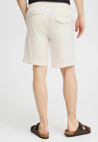 Pantalones cortos beige para hombre con cintura elástica, dos bolsillos traseros y una textura suave. Combinados con sandalias marrones tipo zueco.