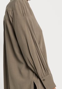Chemise à manches longues, ample, de couleur vert olive atténué. Fabriquée en tissu lisse avec des détails plissés sur les manches et des poignets boutonnés.