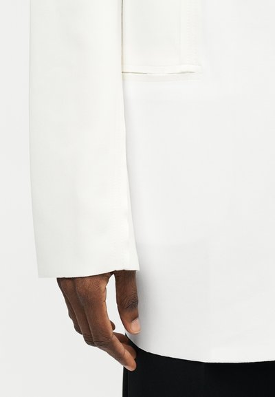 Χέρι με σκουρόχρωμο τόνο δέρματος που ακουμπά κοντά στο τελείωμα ενός λευκού μακρυμάνικου ενδύματος σε ένα ομοιόμορφο ανοιχτό φόντο.