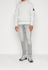 Ljusgrå sweatshirt med framficka och dragsnodd i huvan, parat med ljusgrå jeans och grå sportskor med svarta accenter.