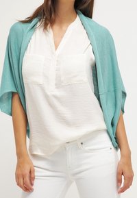 Lätt turkost cardigan över en vit skjorta med två bröstfickor. Bärs med vita byxor som har en avslappnad passform.