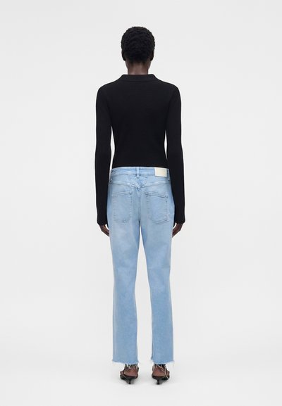 Γυναίκα που στέκεται με την πλάτη γυρισμένη φοράει μαύρο μακρυμάνικο τοπ, ανοιχτό μπλε τζιν με ξεφτισμένα τελειώματα και μαύρες ανοιχτές γόβες σε λευκό φόντο.