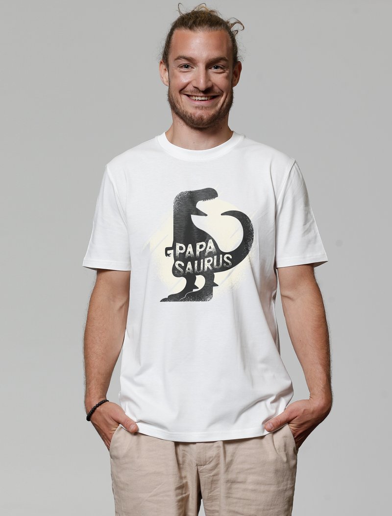 Witte katoenen t-shirt met een zwart dinosaurussymbool en de tekst "Papa Saurus." Korte mouwen en een relaxte pasvorm, gedragen met beige broeken.
