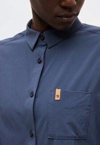 Közelkép egy személyről, aki tengerészkék gombos ingben van, amelyen mellkasi zseb található, és a zsebön egy kis, barna bőrből készült logófül.