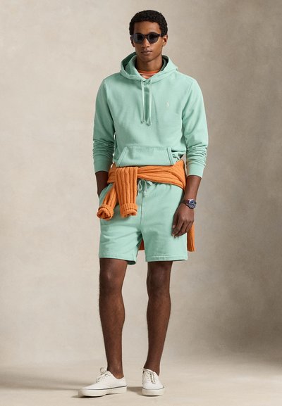 Mátově zelená mikina s kapucí a klokaní kapsou, doplněná o odpovídající šortky. Oranžový pletený svetr uvázaný v pase. Bílé tenisky dokončují outfit.