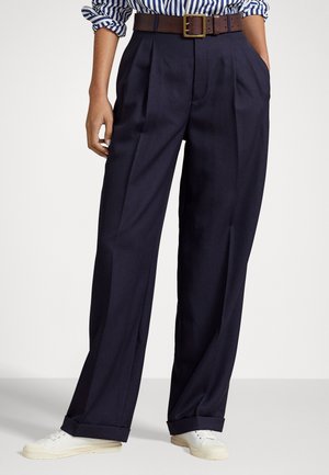 Modèle portant un pantalon large bleu marine taille haute avec revers, ceinture marron, chemise rayée bleue et blanche, et baskets blanches, les mains dans les poches.