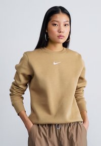 Sweatshirt Nike beige com gola redonda e mangas com punhos. Apresenta um logo swoosh branco no peito. Combinado com calças castanhas claras.