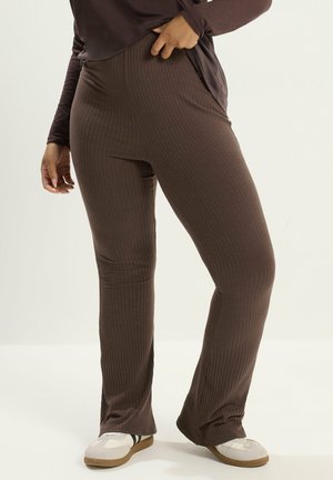 Person iført brune ribstrikkede bukser og matchende langærmet top, stående med den ene hånd på hoften og hvide sneakers.