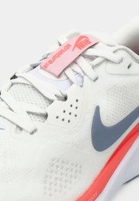 Αθλητικό παπούτσι Nike με λευκό διχτυωτό πάνω μέρος, γκρι λεπτομέρειες, ροζ λουρί και κόκκινη σόλα με υφή.