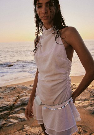 Joven con el pelo mojado, vestida con un vestido blanco sin mangas y cadena decorativa en la cintura, de pie en una playa rocosa al atardecer.