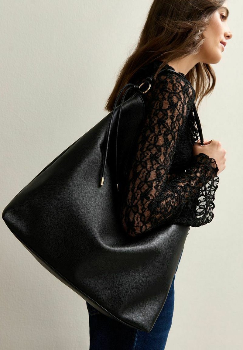 Bolso tote de cuero negro con una textura suave, con cierre de cordón y acentos en tono dorado. Diseñado para la practicidad y el estilo.