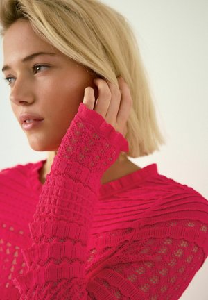 Fuksjowy sweter dziany z teksturowanymi paskami i koronkowym wzorem, z ażurowymi rękawami i prążkowanym kołnierzem. Zbliżenie na szczegóły projektu.