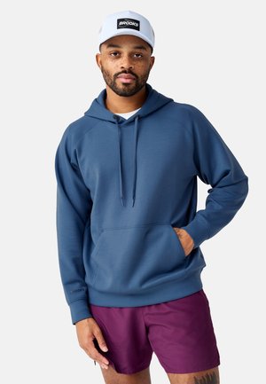 Hombre con barba que lleva una sudadera con capucha azul, gorra blanca con la palabra "Brooks" y pantalones cortos morados, con una mano en el bolsillo de la sudadera, mirando hacia adelante.