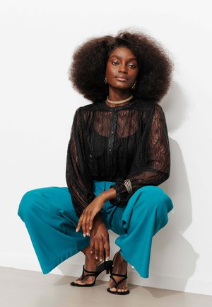 Femme aux cheveux bouclés volumineux portant un chemisier en dentelle noire, un pantalon large bleu sarcelle, des sandales noires à talons et des bijoux en or, accroupie contre un mur blanc.