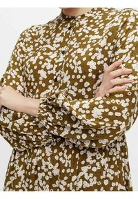 Kvinna med armarna i kors, iklädd en brun långärmad klänning med ett vitt och rosa blommönster samt svarta knappar vid kragen.