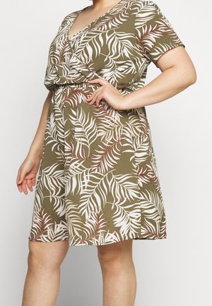 Kobieta w krótkim, sięgającym kolan oliwkowozielonym sukience z krótkim rękawem, ozdobionej białymi i brązowymi liściastymi wzorami, z ręką spoczywającą na talii.