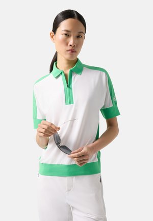Woman with straight black hair in a white and green short-sleeve polo shirt holding black sunglasses against a plain light background.