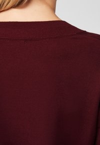 Rückansicht einer Person in einem bordeauxroten Strickpullover, der den Halsausschnitt und den oberen Rücken mit heller Haut und blondem Haar zeigt.