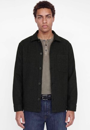 Jeune homme aux cheveux bouclés portant une veste sombre boutonnée par-dessus un t-shirt henley beige et un jean bleu sur un fond blanc uni.