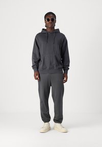 Grau Baumwollhoodie mit Kordeln und Kängurutasche, kombiniert mit grauen Jogginghosen und hellen Sneakers. Person trägt Sonnenbrille.