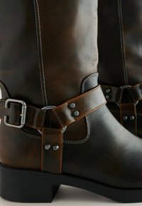 Brązowe skórzane kozaki do kolan z postarzanym wykończeniem, z szerokim paskiem, metalowymi klamrami i blokowym obcasem.