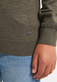 Maglione lavorato a maglia verde oliva con polsini e orlo a coste, caratterizzato da una piccola applicazione metallica del logo. Abbinato a jeans blu chiaro.