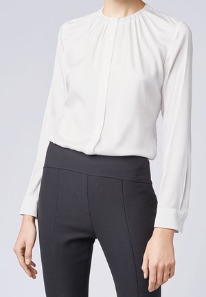 Nainen, joka pukeutunut valkoiseen pitkähihaiseen paitapuseroon, jossa on laskoskaulus, ja mustiin korkeavyötäröisiin, istuviin housuihin, seisoo yksivärisen taustan edessä.