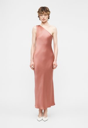 Frau steht frontal, trägt ein ärmelloses, einseitig schulterfreies, bodenlanges Kleid in staubigem Rosaton und weiße spitze Schuhe vor einem schlichten Hintergrund.