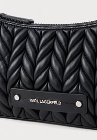 Fekete bőr táska kiemelkedő, csavart mintával. Ezüst színű kiegészítővel és merész logótáblával rendelkezik. Cipzárral zárható felül.