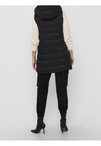 Person, der einen langen schwarzen gepolsterten Weste mit Kapuze, einen cremefarbenen Strickpullover, schwarze Cargo-Hosen und schwarze, mit Absatz versehenen Ankle-Boots trägt, steht mit dem Rücken zu uns.